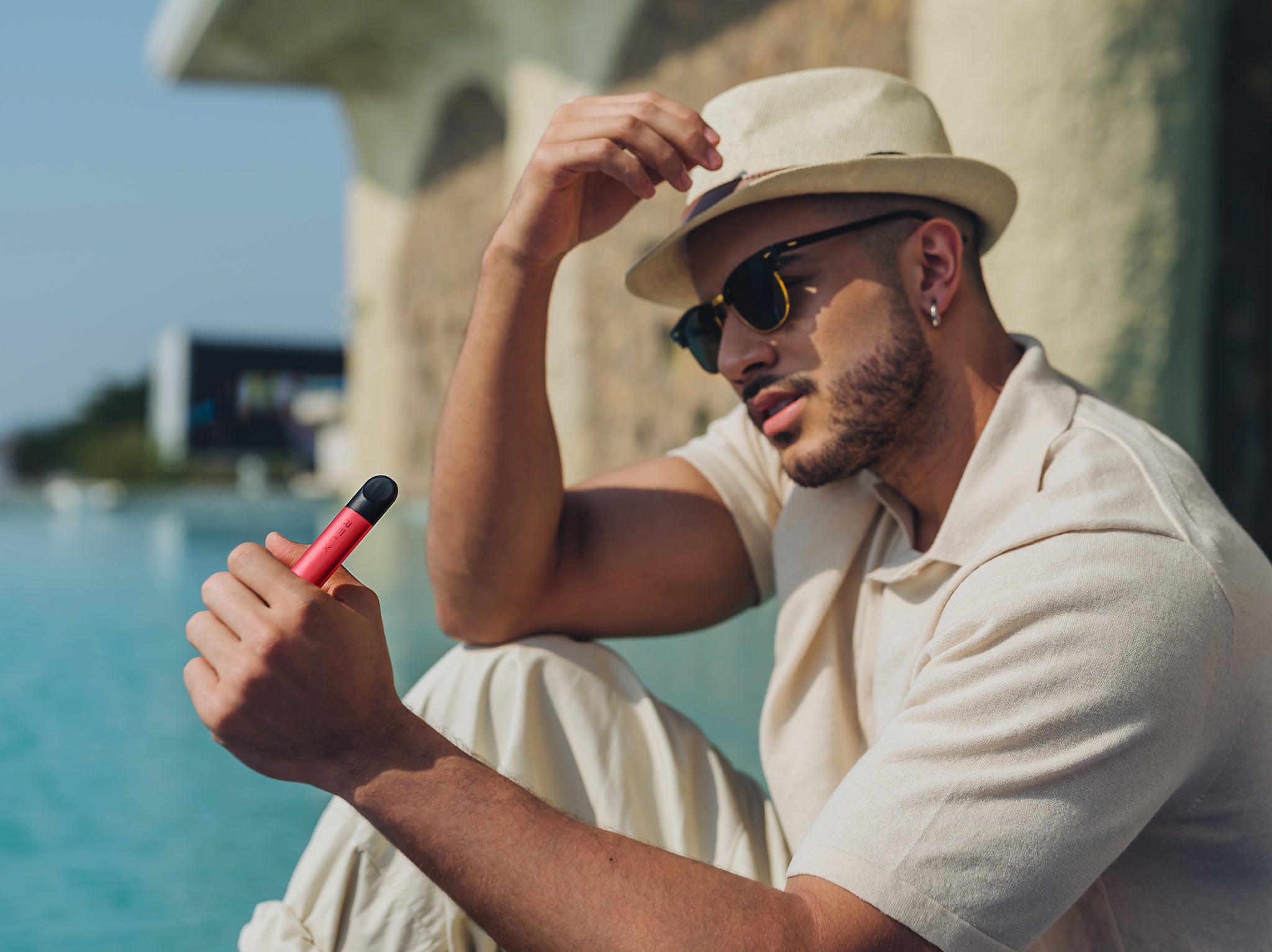 Young bearded man vaping red RELX Infinity and staring into the sun as he sits besides the sea