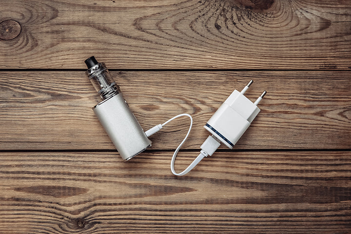 A silver vape and white charger lie on a wooden table.